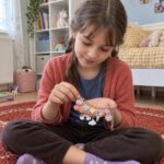 Момиче седи на килима и разглежда гривна с цветни висулки в дланите си.