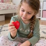 Момиче с плитка седи на килима и стиска червеното сърце в ръка.