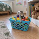 Синята детска кошница с храни на дървения под в детската стая, до плюшено мече и рафт с книги.