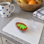 Отворено яйце с оранжев слайм и динозавърче върху бяла подложка на кухненски плот до чаша и купа с плодове.