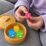 Детски пръсти държат розовото кубче с брокат, а синьото, жълтото и зеленото чакат в отворената кутийка на сивия диван.