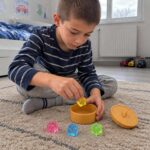 Момче седи на килима и прибира жълтото скуиши кубче в отворената кутийка.
