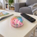 Розово-дъговата кутийка putty slime с еднорог на холна маса до дистанционно и отворена книга.