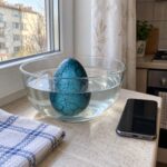 Синьото динозавърско яйце във вода в стъклена купа на кухненски плот до телефон и кърпа.