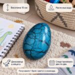 Синьо динозавърско яйце на килим до тетрадка с рисунка, с надписи „Височина 10 см“, „Расте във вода“, „Динозавър в яйцето“ и „Следи промяната ден по ден“; продава се 1 брой с цвят изненада.