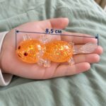 Оранжев прозрачен аксолотъл с брокат в отворена длан, със стрелка за дължина 8,5 см.