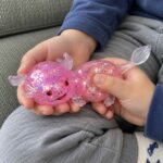Детски ръце държат розовия разтеглив аксолотъл и натискат с палец гърба му.