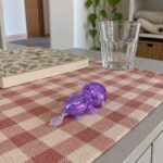 Лилав разтеглив аксолотъл с брокат на маса с карирана покривка до празна чаша.