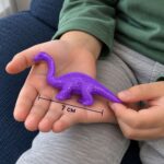 Лилаво дълговрато динозавърче в детска длан, с линия за дължина 7 см.