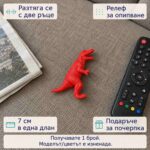 Червено разтегливо динозавърче на дивана до дистанционно, с надписи за разтягане с две ръце, релеф за опипване и 7 см в длан; продава се 1 брой с модел и цвят изненада.