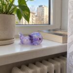 Лилав скуиши аксолотъл на перваза на прозореца до книга и саксия с растение.