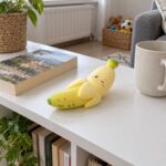 Скуиши банан с лице, легнал на бяла масичка до книга и чаша.