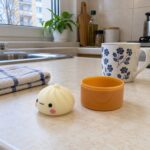 Бял скуиши дъмплинг на кухненски плот до оранжевата кутийка и чаша на цветя.