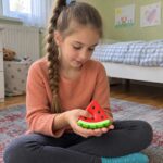 Момиче с плитка седи на килима и държи скуиши динята в двете си длани.