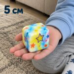 Синьо-жълто скуиши кубче със звезди в отворена детска длан над килима, с надпис 5 см.