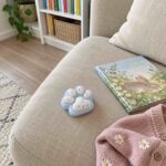 Светлосиня скуиши лапичка на бежов диван до книжка със зайче и розова плетена жилетка.