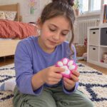 Момиче с кок седи на килима и натиска с пръсти розовата скуиши лапичка.