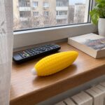 Скуиши царевица на дървен перваз до дистанционно и книга, с изглед към блоковете навън.