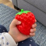 Детска ръка стиска червената скуиши ягода над синия килим.