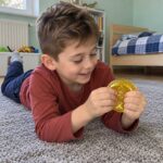 Момче лежи по корем на килима и стиска с две ръце златната скуиши монета.