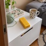 Скуиши златно кюлче на бяло шкафче до дивана, редом с кълбо зелена прежда, книга и чаша.