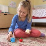Момиче седи на килима и подрежда синьо кученце и ягода, а в дланта си държи овчица.