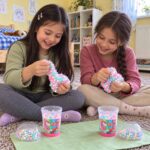 Две момичета мачкат слайм с пастелни топчета пред две чаши Super Slime и капаците им.